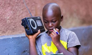 A young boy in Rwanda, identified as Igihozo Kevin, 11, listens to his Primary 5 lessons on a radio while studying at home due to school closures caused by the coronavirus pandemic. He is holding a radio and a pen, focused on his notebook.