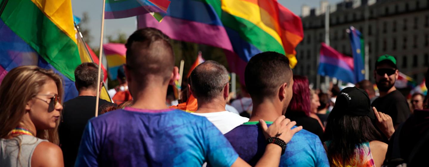 Un couple de même sexe avec les bras autour des épaules l'un de l'autre lors d'une marche fierté, avec des drapeaux arc-en-ciel en arrière-plan.