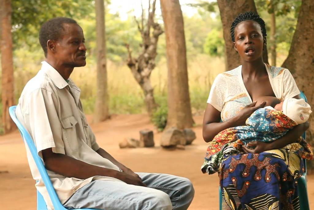 Mwanamume Mtanzania ameketi kwenye kiti cha bluu karibu na mwanamke anayemnyonyesha mtoto wake katika kliniki ya nje huko Sengerema, Tanzania.