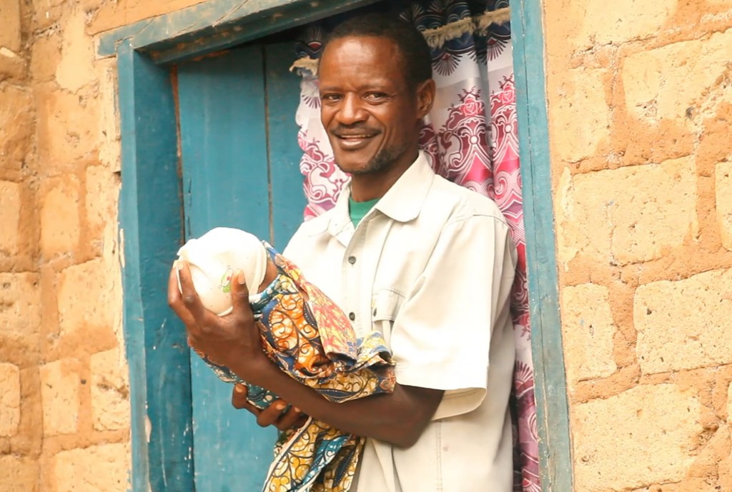 Mwanamume mwenye tabasamu aliyevaa shati jeupe amemshika mtoto mchanga aliyefunikwa kwa blanketi lenye rangi nyingi, amesimama mlangoni.