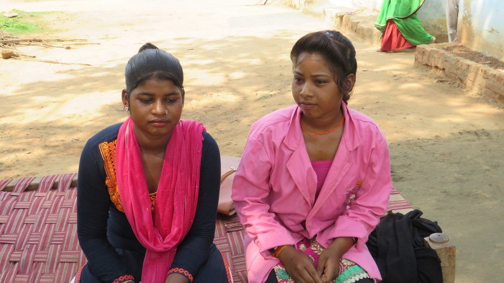 भारत में दो युवतियां, लाली आदिवासी और रामकली आदिवासी, बाहर एक बुनी हुई चटाई पर बैठी हैं।