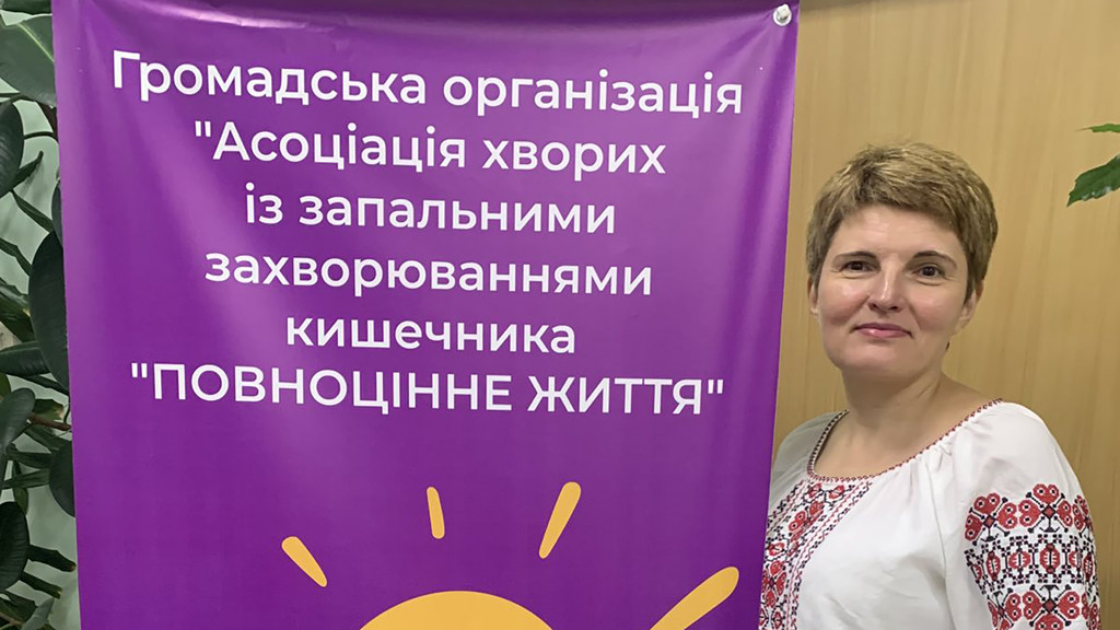 Украинка Елена Соцкова стоит рядом с фиолетовым баннером ассоциации пациентов с воспалительными заболеваниями кишечника.