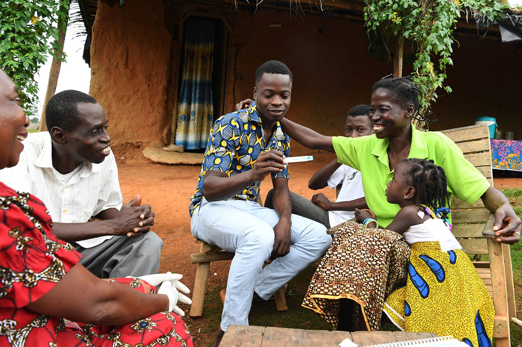 Familia ya Kiafrika imeketi pamoja nje ya nyumba huko Benjaminkro, Côte d'Ivoire, huku baba akimfanyia mtoto wake kipimo cha VVU nyumbani.