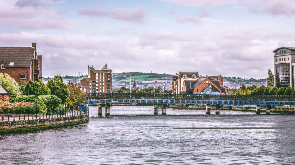 Una vista de Belfast con un puente azul sobre el río Lagan, flanqueado por edificios modernos e históricos bajo un cielo nublado.