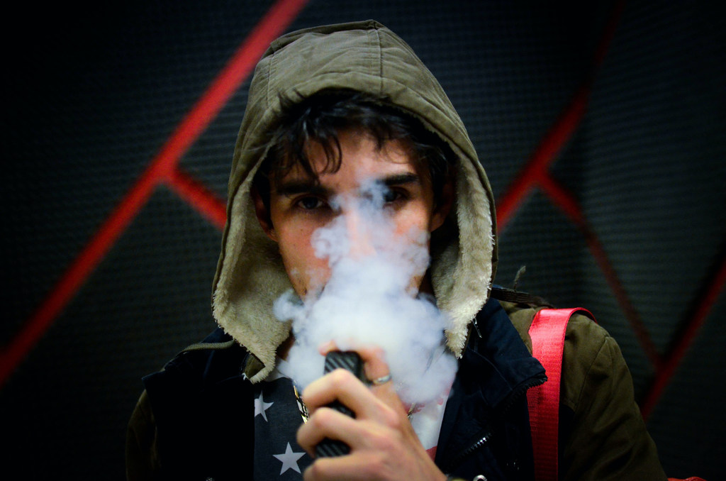 Un jeune homme portant une veste à capuche exhale un important nuage de vapeur provenant d'une cigarette électronique.