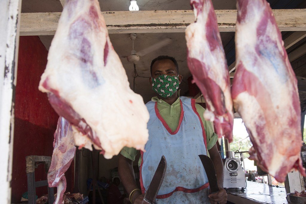 Mchinjaji aliyevaa barakoa ya kitambaa chenye muundo amesimama nyuma ya kibanda chake sokoni, huku vipande vikubwa vya nyama vikining'inia mbele.
