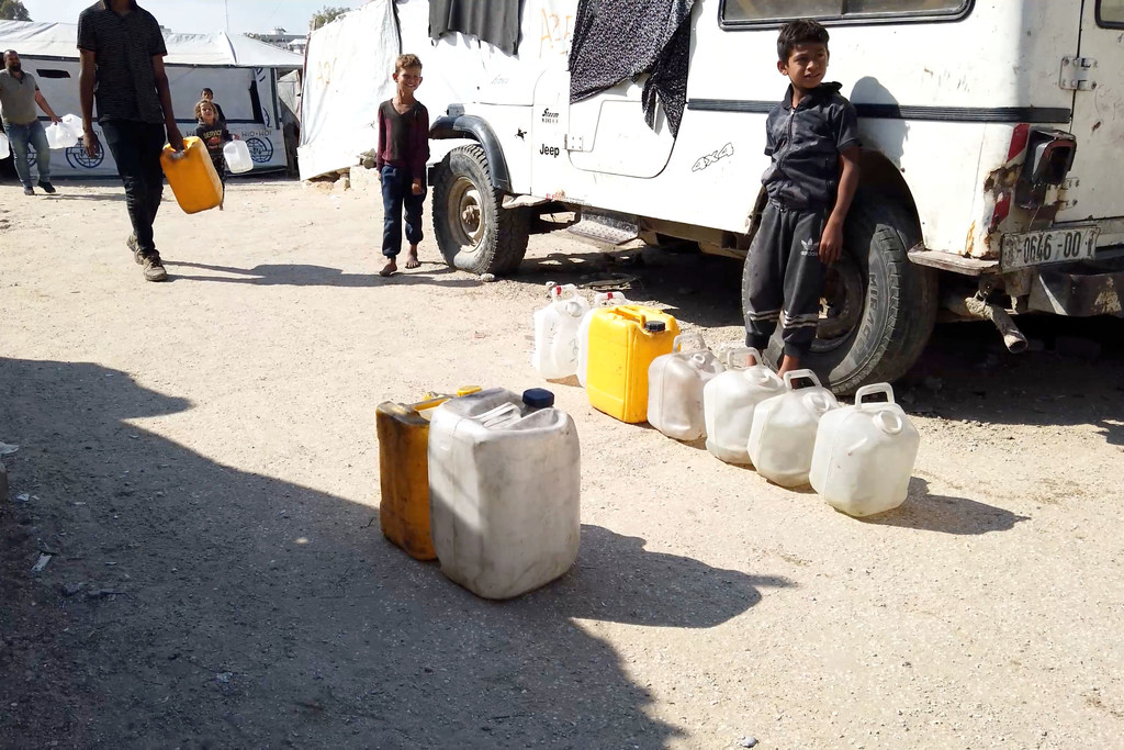 أطفال وكبار يملؤون حاويات بلاستيكية بالماء من شاحنة صهريج بيضاء في مخيم للنازحين في غزة.