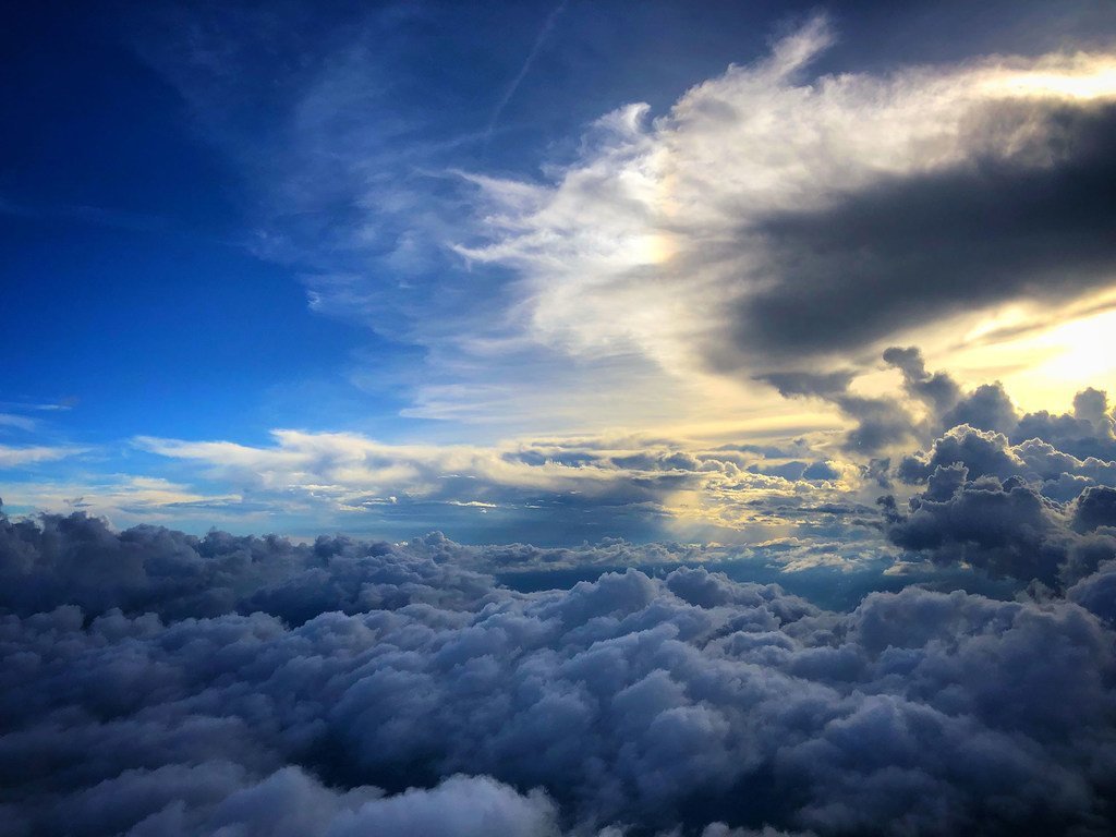 Mtazamo kutoka juu wa safu nene ya mawingu ya cumulus, ukiangalia juu angani ya bluu huku miale ya jua ikipita kwenye mawingu upande wa kulia.