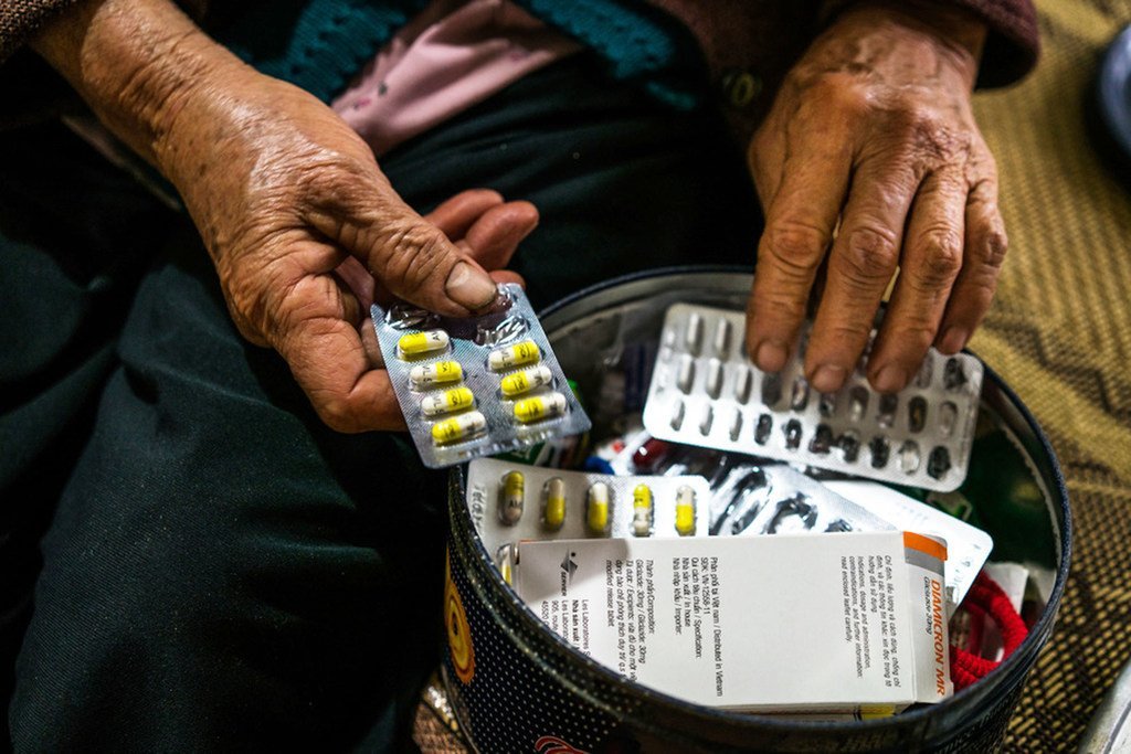 Um close das mãos de uma senhora idosa segurando uma cartela de comprimidos sobre um recipiente com outros medicamentos.