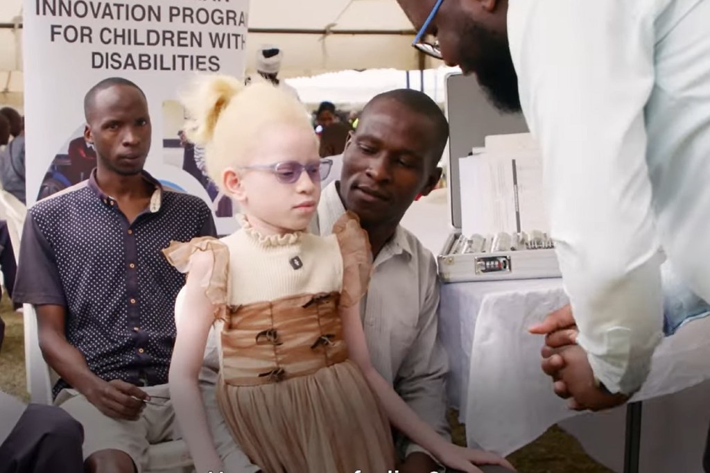 Mtoto mwenye umri wa miaka 7 mwenye ulemavu wa ngozi, anayeitwa Candy, ameketi kwenye hafla nchini Kenya kwa ajili ya watoto wenye ulemavu, kama sehemu ya mpango wa UNICEF . Amevaa gauni la beige na kahawia na miwani ya jua, huku wanaume wawili wakiwa wamesimama kando yake.