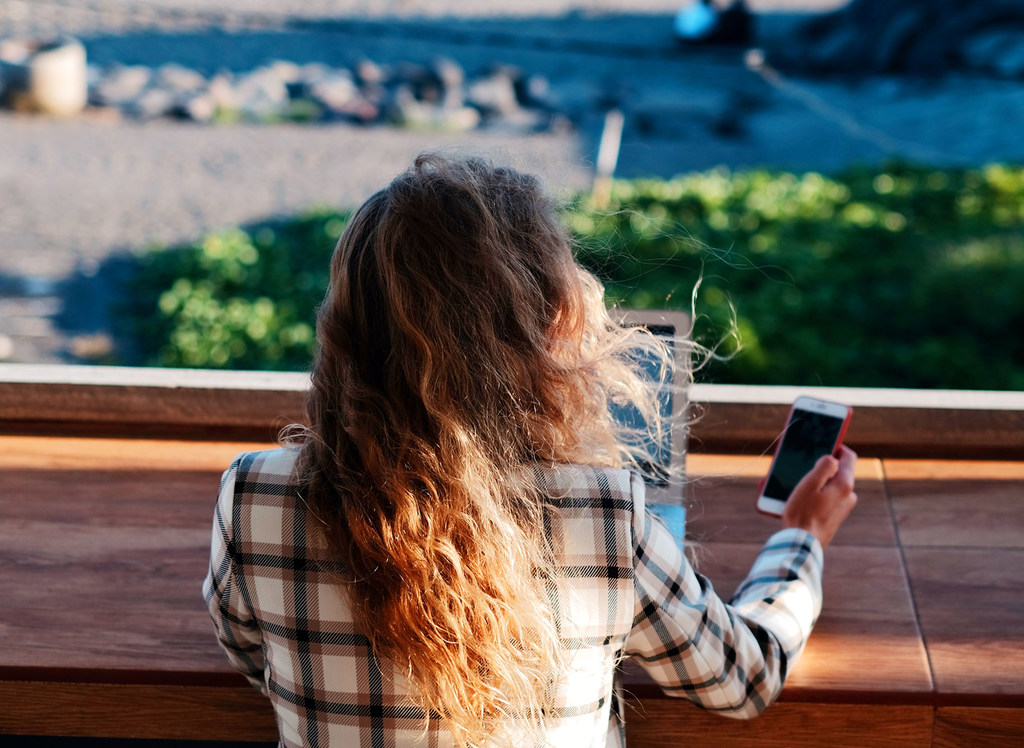 Una mujer de cabello largo y rizado, vista de espaldas, con una camisa a cuadros, está sentada en un banco de madera con una computadora portátil y un teléfono inteligente. Contempla un paisaje de vegetación y agua.