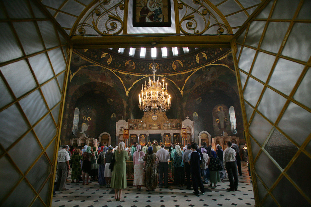 Interior view of a church in Kyiv-Pechersk Lavra, showing a congregation attending Sunday mass with a large chandelier and religious icons.