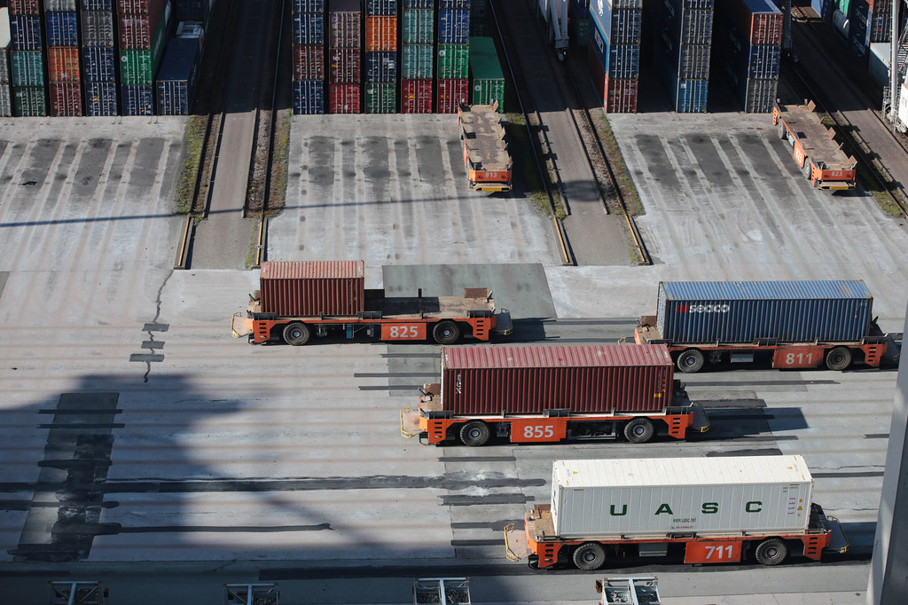 Vista aérea de um terminal de contêineres mostrando várias máquinas de movimentação de contêineres, vestindo roupas laranja, transportando contêineres por um pátio pavimentado.