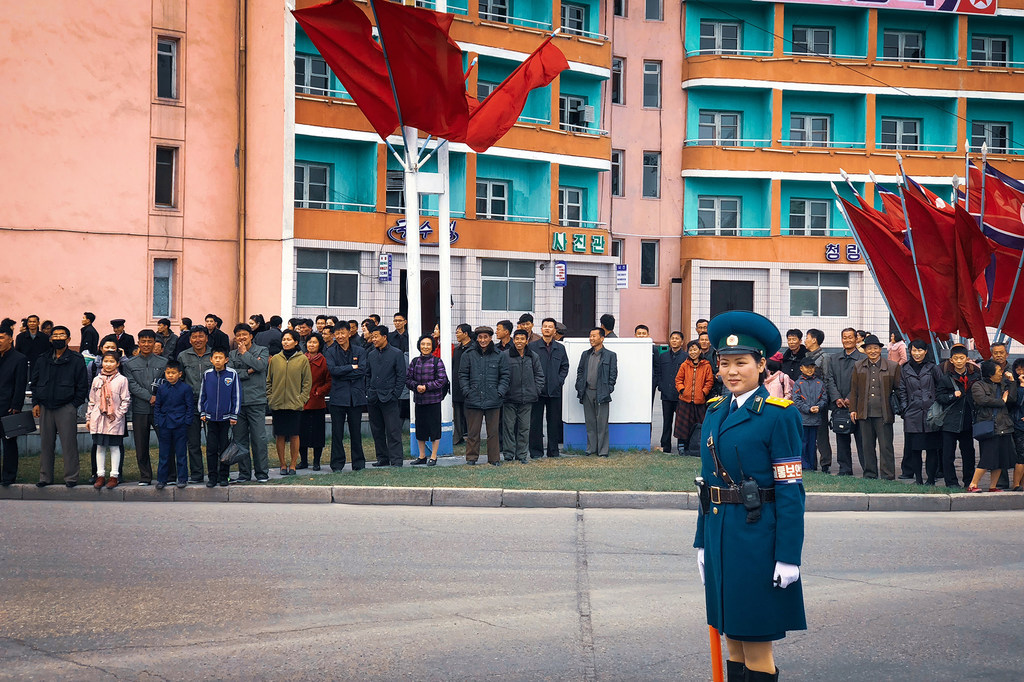 Un guardia uniformado permanece firme en una calle de Corea del Norte, con una multitud de civiles y banderas rojas al fondo.