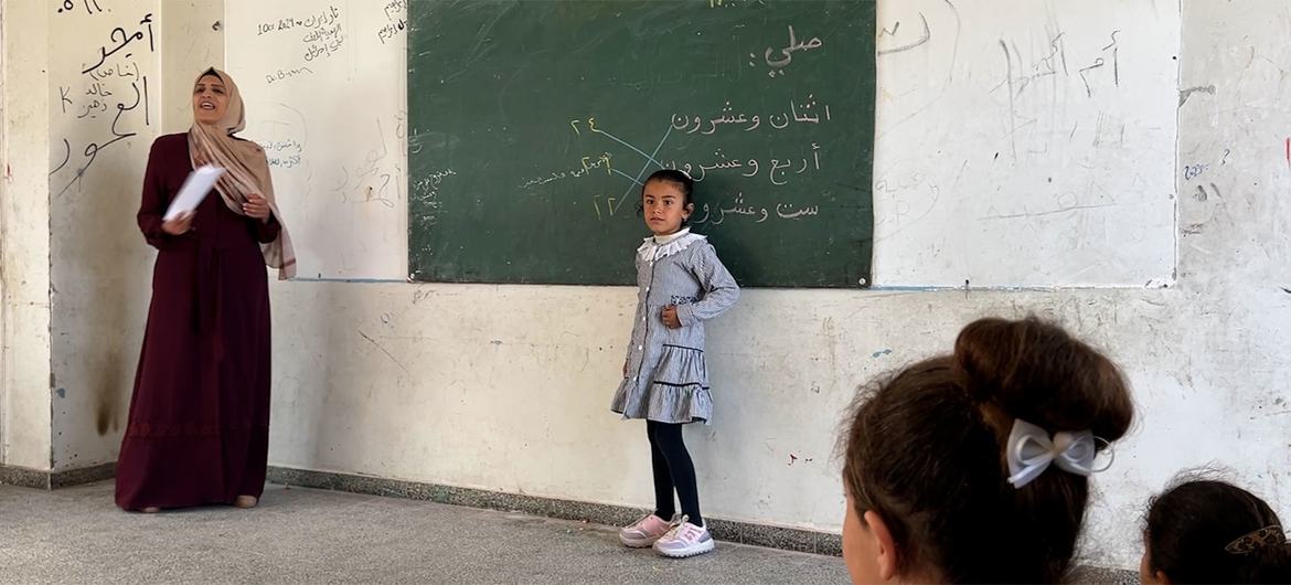 تقف معلمة فلسطينية ترتدي الحجاب والزي التقليدي في فصل دراسي تزين جدرانه كتابة عربية، ويوجد سبورة. تقف بالقرب منها تلميذة فلسطينية صغيرة ترتدي الزي المدرسي، بينما يجلس طلاب آخرون في المقدمة.