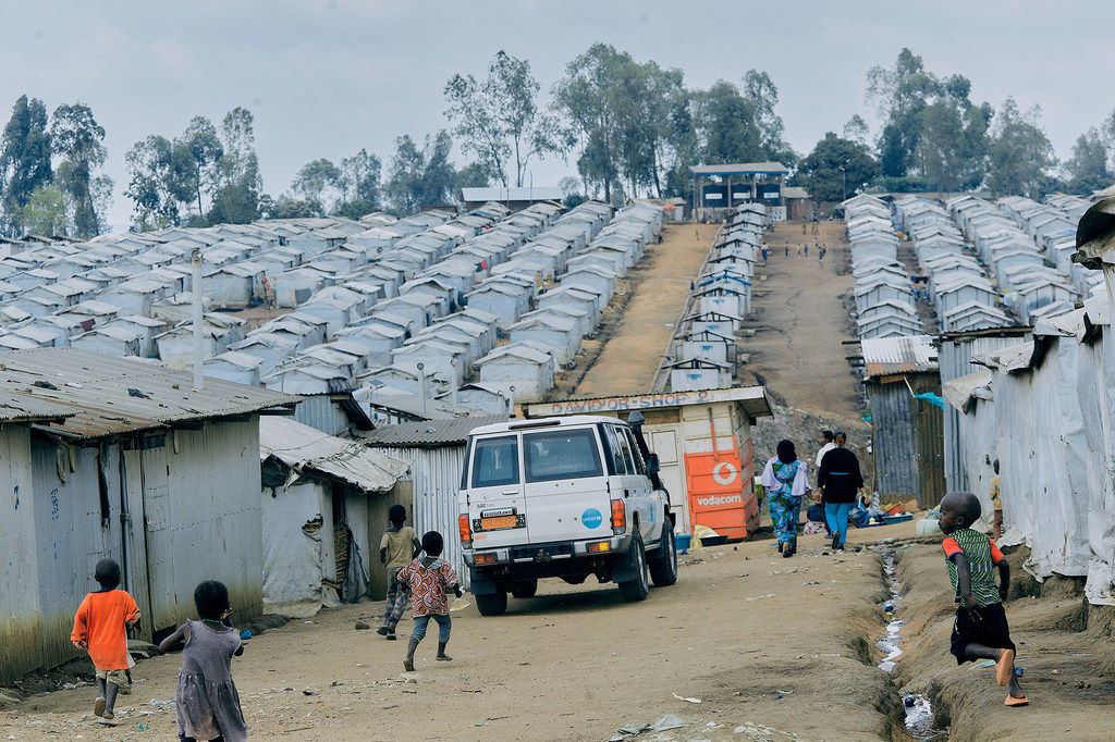 Des enfants jouent sur un chemin de terre dans un grand camp de déplacés, avec des rangées d'abris de fortune en arrière-plan, et un véhicule d'aide blanc au centre.