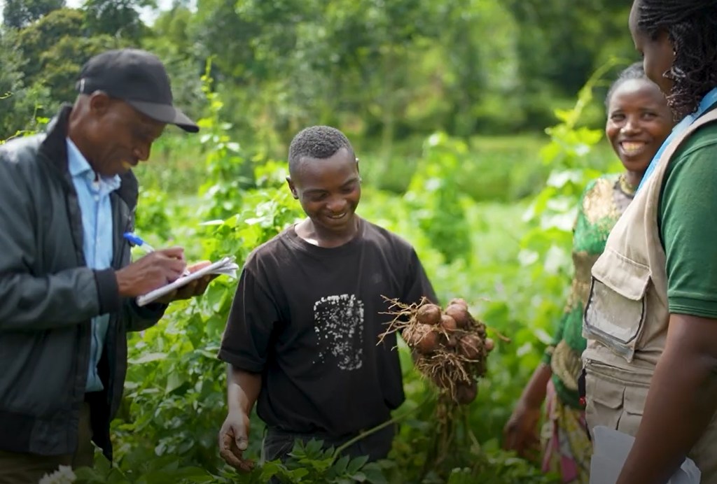 Mwanamume akiwaonyesha kikundi cha wafanyakazi wa kilimo viazi vilivyovunwa shambani nchini Rwanda, huku mwakilishi wa FAO akiandika maelezo.