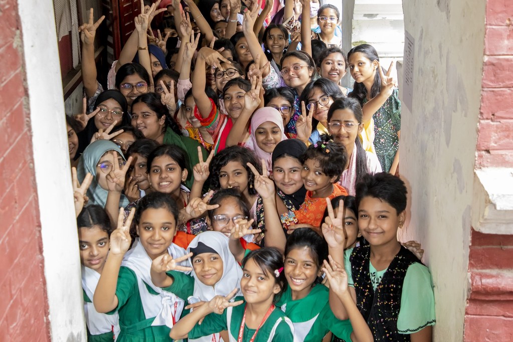 बांग्लादेश के एक स्कूल में मुस्कुराती हुई दक्षिण एशियाई लड़कियों का एक समूह टीकाकरण के लिए कतार में इंतजार करते हुए शांति का संकेत दे रहा है।