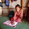 एक युवा रोहिंग्या लड़की अपने घर में एक अस्थायी आश्रय में चटाई पर बैठकर पढ़ाई कर रही है, उसके पास UNICEF के बैग और एक बैकपैक पड़ा है, क्योंकि उसका शिक्षण केंद्र बंद है।