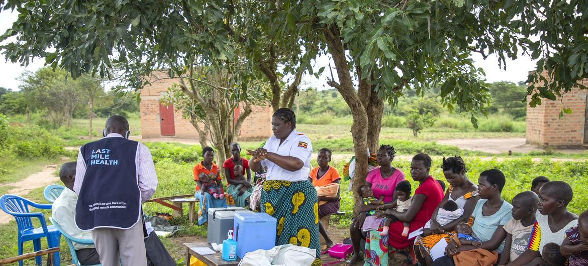 Mhudumu wa afya na mkunga wakijiandaa kwa ajili ya huduma ya chanjo katika Kijiji cha Lwalu, Uganda, huku umati wa akina mama na watoto wakisubiri karibu.