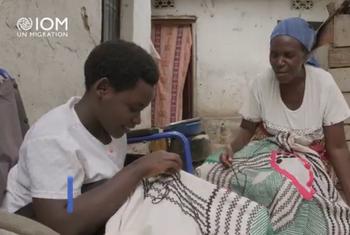 Mwanamke kijana na mwanamke mzee wanashona shuka pamoja. Nembo ya IOM inaonekana kwenye kona ya juu kushoto.