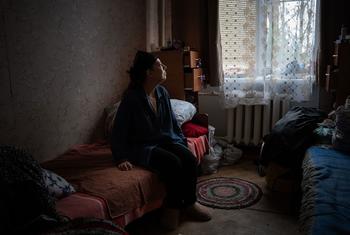 A woman sits alone on a bed in a dimly lit room, looking upwards in apparent grief. She is from the Horovan district in Kherson, Ukraine, and has lost both her husband and mother to war-related incidents.