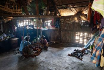 Inside a makeshift bamboo shelter in an IDP camp in Rakhine State, Myanmar, showing a sleeping baby in a woven basket, a mother, and another child, with clothes hanging and sunlight filtering through the walls.