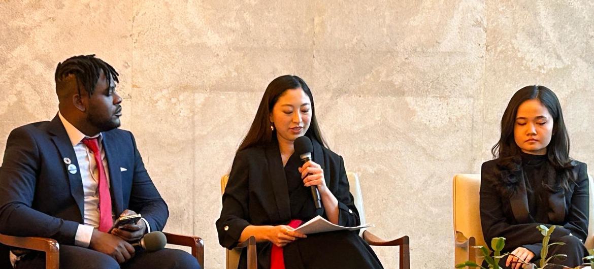 امرأة ترتدي سترة سوداء تتحدث إلى الميكروفون وهي تحمل أوراقًا، جالسة بين رجل أسود يرتدي بدلة وامرأة أخرى في مناسبة رسمية.