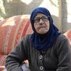 Uma senhora palestina idosa, com um lenço azul na cabeça e óculos, está sentada ao ar livre em uma cadeira estampada.