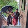 बांग्लादेश में बारिश के दौरान दो महिलाएं और एक बच्चा नए ईंट के घर की सीढ़ियों से नीचे उतर रहे हैं। एक महिला ने बच्चे के ऊपर छाता पकड़ रखा है।