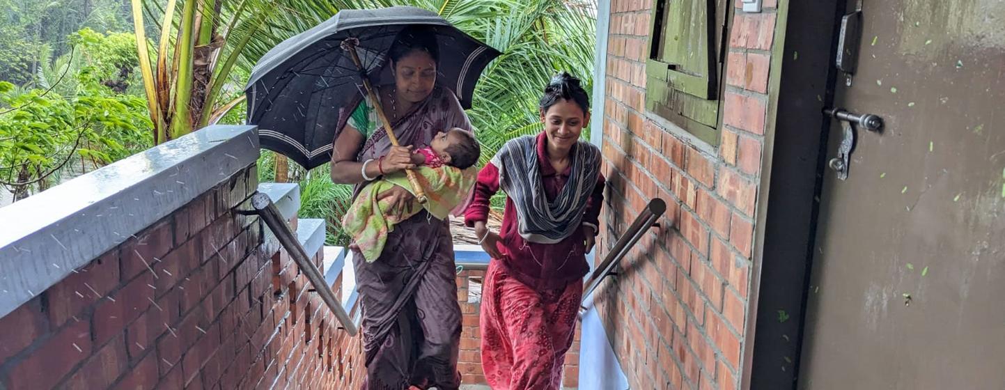 बांग्लादेश में बारिश के दौरान दो महिलाएं और एक बच्चा नए ईंट के घर की सीढ़ियों से नीचे उतर रहे हैं। एक महिला ने बच्चे के ऊपर छाता पकड़ रखा है।
