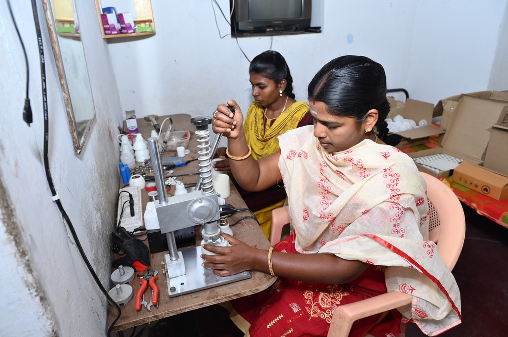 डी पूविझी एक कार्यशाला में मशीन चला रही हैं, जबकि एक अन्य महिला उन्हें देख रही है।