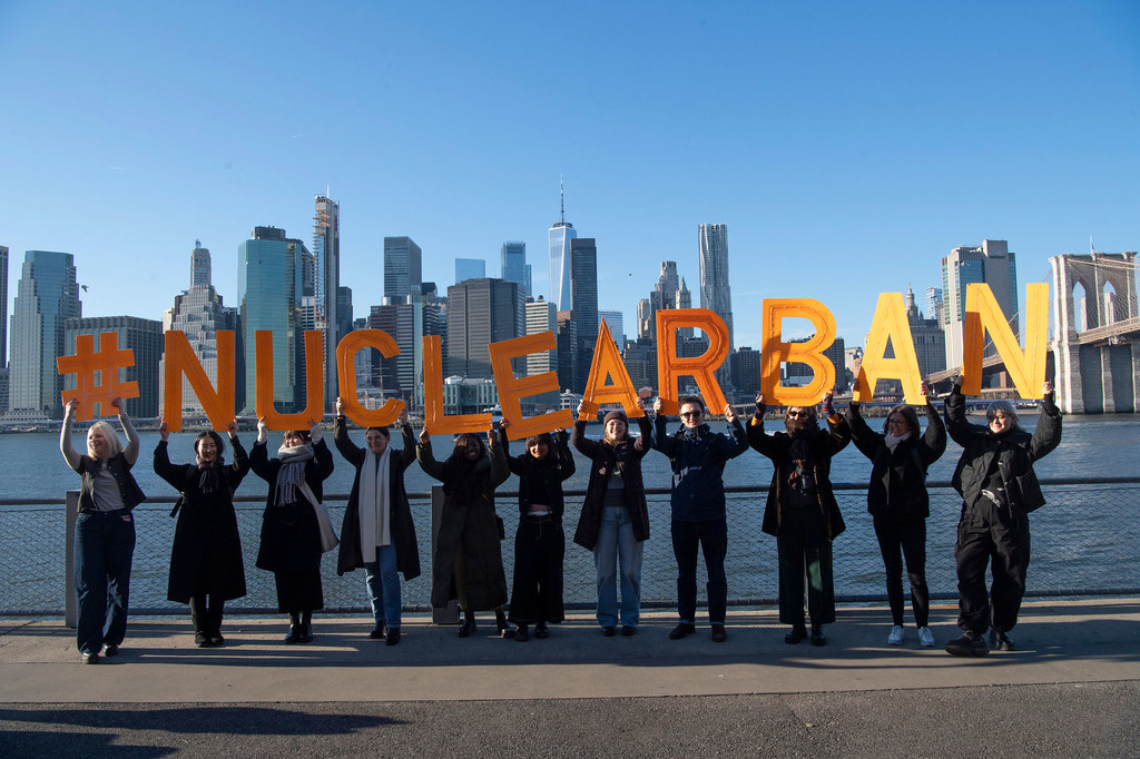 يرفع ناشطو منظمة "إيكان" حروفًا برتقالية كبيرة تُشكّل كلمة #NUCLEARBAN، مع ظهور أفق مدينة نيويورك وجسر بروكلين في الخلفية.