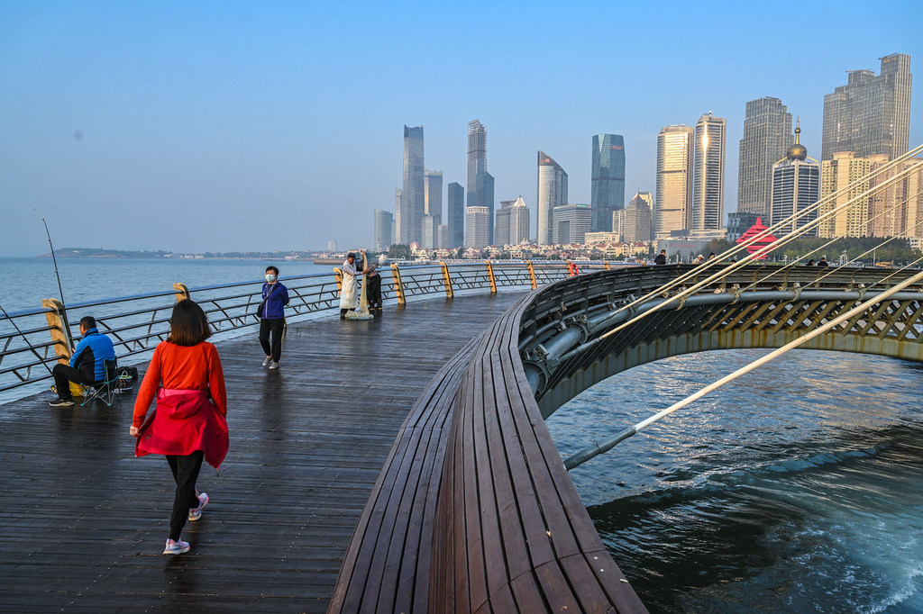La gente pasea y pesca en un paseo marítimo de madera en la Plaza del Cuatro de Mayo en Qingdao, con el moderno perfil urbano de la ciudad de fondo.