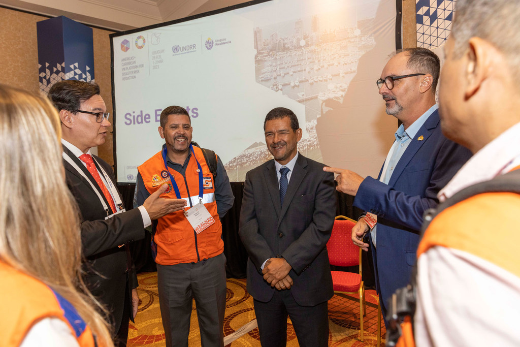 Raúl Salazar y otros hombres vestidos de manera profesional participan en una conversación durante un evento paralelo a la conferencia. Al fondo se observa una pantalla de presentación.