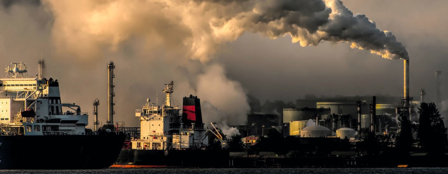 A view of a polluted industrial waterfront at sunset, with large plumes of dark smoke billowing from factory smokestacks into the sky. Several large ships are docked in the foreground on the water.