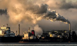 A view of a polluted industrial waterfront at sunset, with large plumes of dark smoke billowing from factory smokestacks into the sky. Several large ships are docked in the foreground on the water.