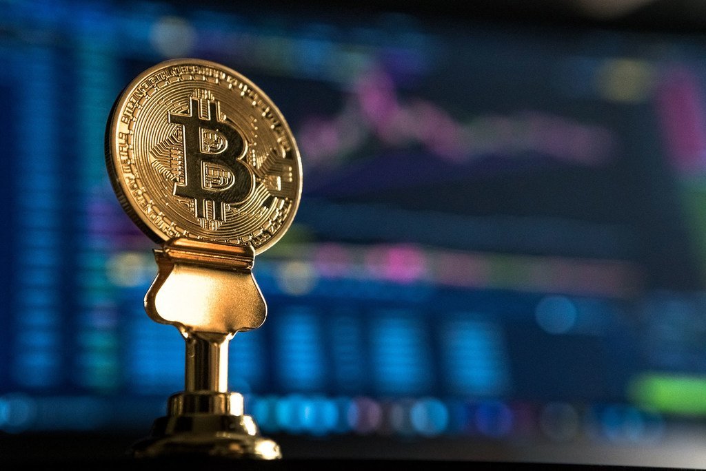Una moneda física de Bitcoin sobre un soporte frente a una pantalla que muestra gráficos financieros.