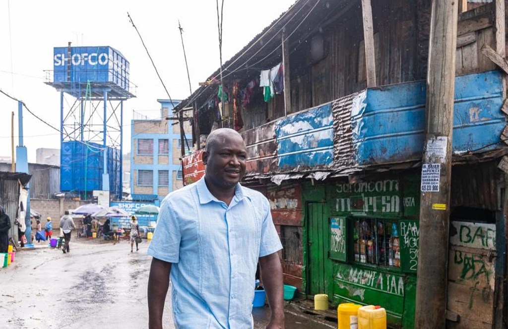 Mwanamume anatembea barabarani huko Kibera, Kenya, akiwa na tanki kubwa la maji la bluu nyuma.