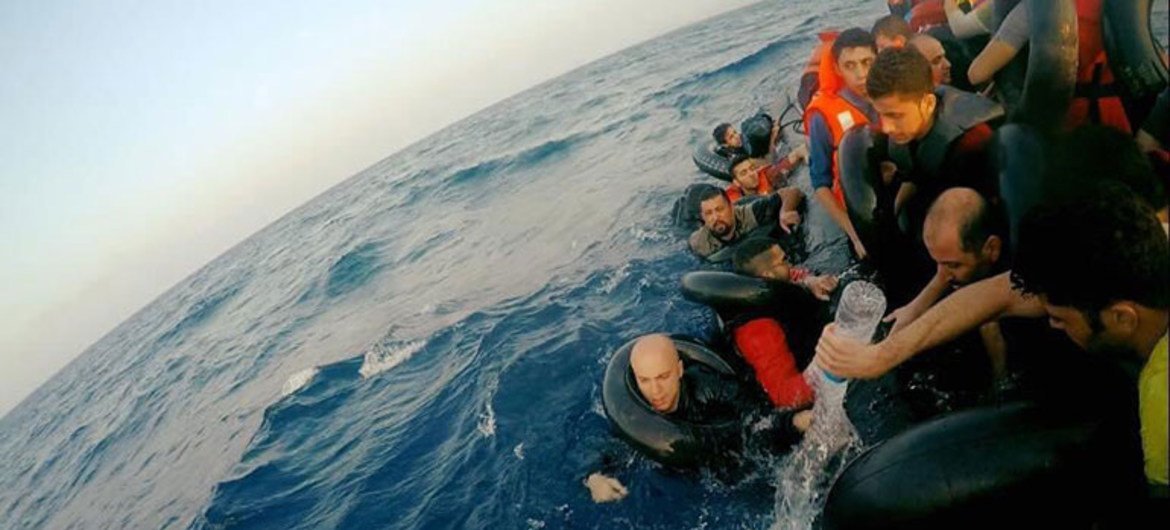A group of refugees on an overcrowded inflatable boat in the Mediterranean Sea, with one person in the water being handed water by a man on the boat.