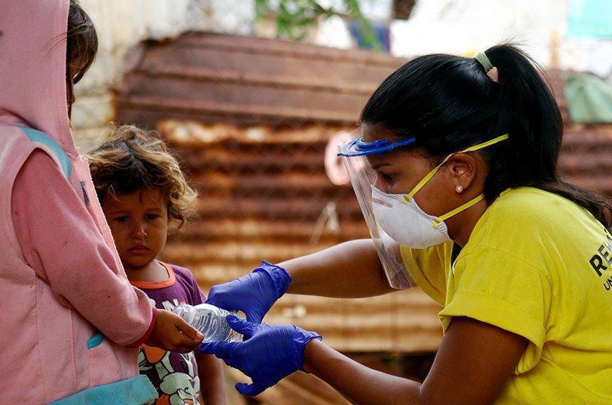 Una trabajadora sanitaria, vestida con una camiseta amarilla, una careta protectora y una mascarilla, distribuye agua a los niños en un espacio al aire libre.