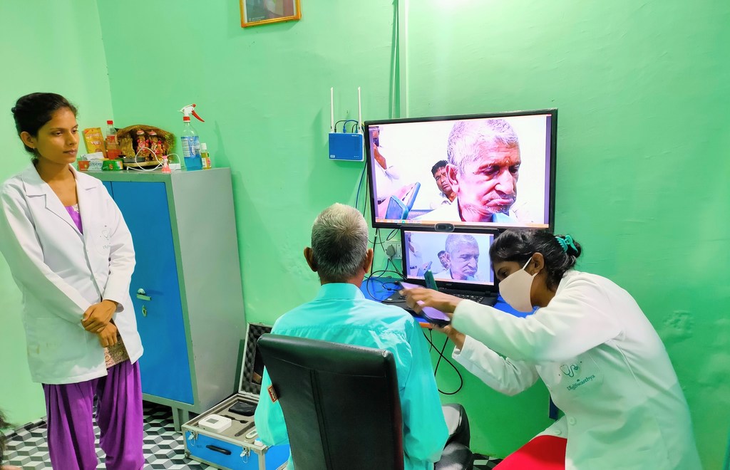 एक क्लिनिक में वीडियो कॉल के माध्यम से स्वास्थ्य परामर्श हो रहा है। एक डॉक्टर और एक नर्स एक मरीज की देखभाल कर रहे हैं, डॉक्टर एक बड़ी स्क्रीन पर दूरस्थ कनेक्शन को सुगम बनाने के लिए टैबलेट का उपयोग कर रहे हैं।