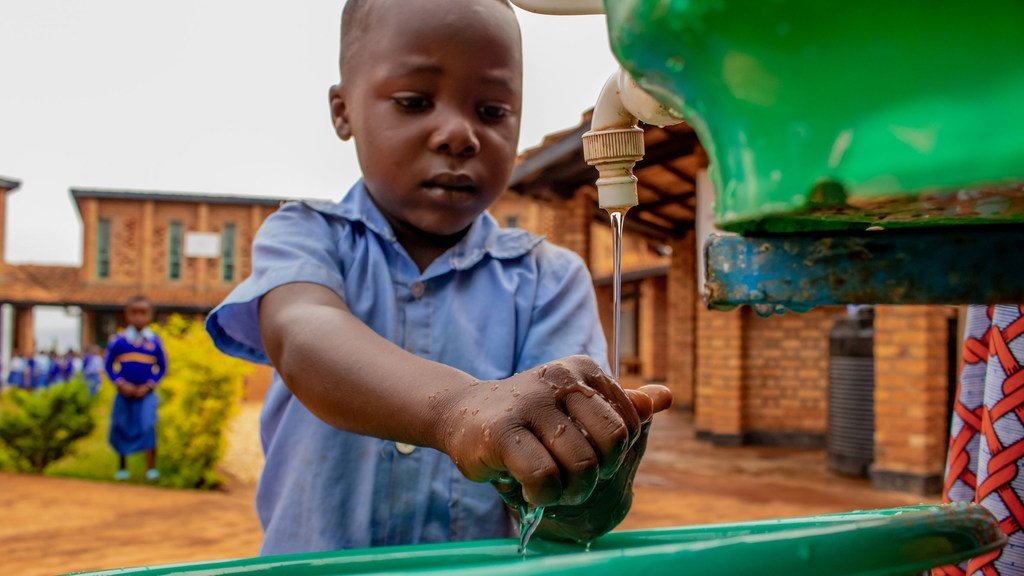 Mtoto mchanga mweusi aliyevaa sare ya shule ya bluu anaosha mikono yake kwenye bomba la maji la kijani katika Kituo cha Maendeleo ya Watoto Wadogo cha Gikomero nchini Rwanda.