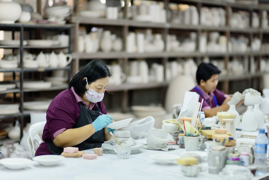 Duas mulheres usando máscaras e luvas pintam peças de cerâmica em uma mesa dentro de uma fábrica de cerâmica.
