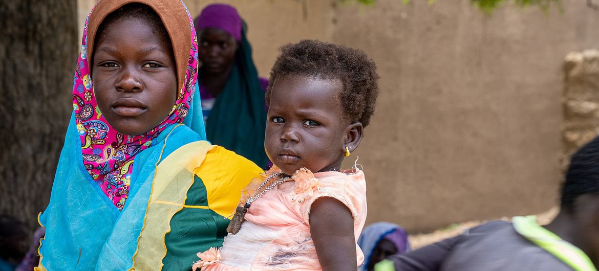 Une femme portant un foulard coloré tient un jeune enfant tandis que d'autres femmes se rassemblent en arrière-plan au Burkina Faso.