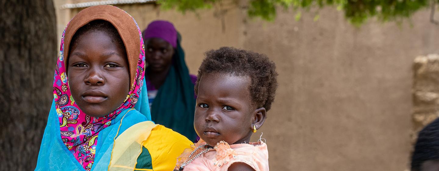 Une femme portant un foulard coloré tient un jeune enfant tandis que d'autres femmes se rassemblent en arrière-plan au Burkina Faso.
