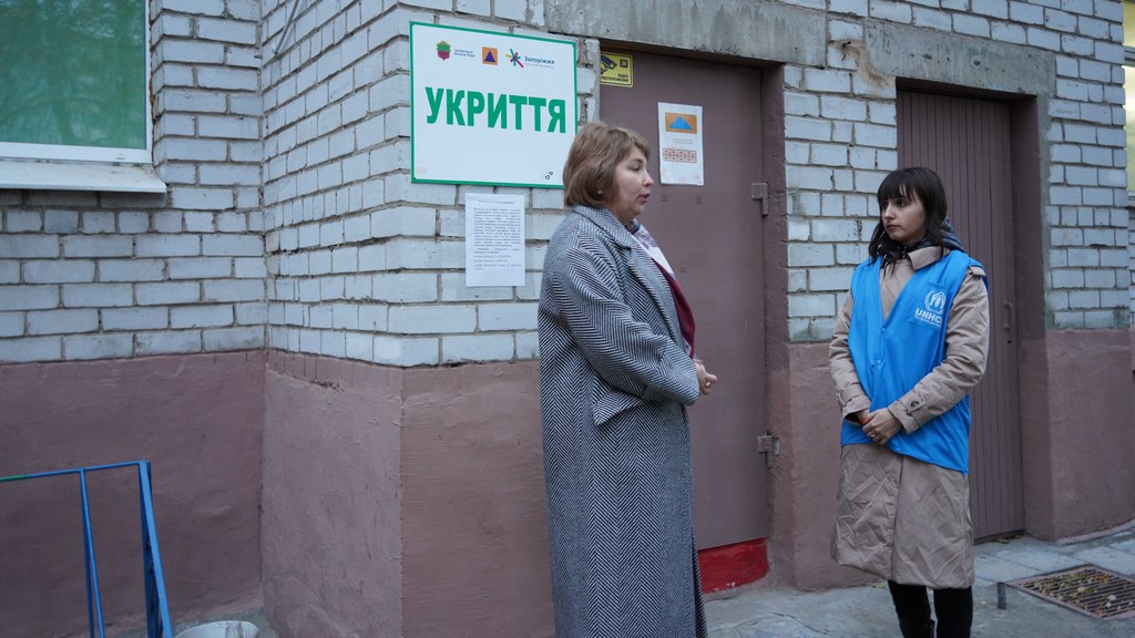 Женщина в синем жилете UNHCR стоит у здания с украинской вывеской приюта и разговаривает с другой женщиной.