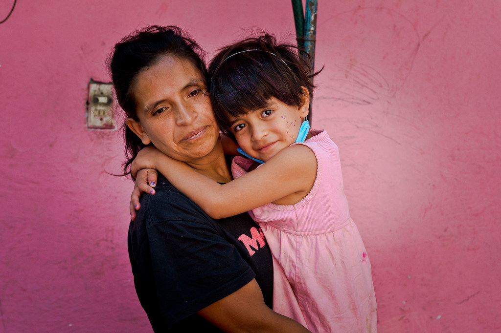 Una mujer y una niña se abrazan frente a una pared rosa.