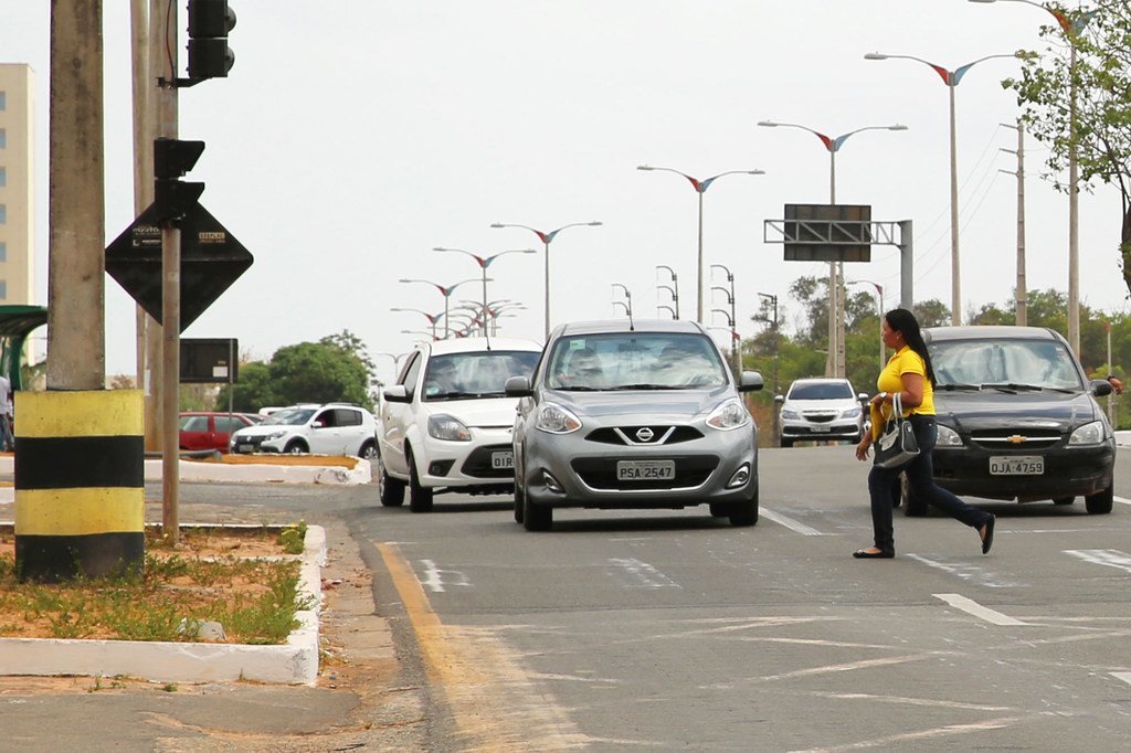 Uma mulher de camisa amarela atravessa uma avenida de várias faixas no Brasil, com diversos carros parados no trânsito atrás dela.