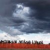 Beneficiários aguardam em uma longa fila atrás de uma cerca de segurança laranja em um ponto de distribuição de alimentos em Malakal, Sudão do Sul, sob um céu escuro e tempestuoso.